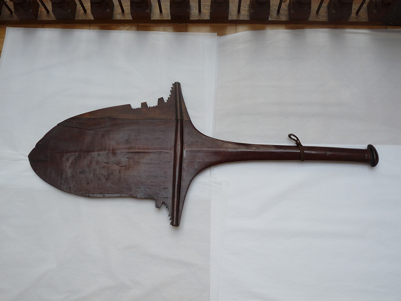 A dark brown wooden paddle, with a large flat head and rounded shaft. Rounded to a point at the end, it has no decoration but does have shaped cuts in both sides near the base.