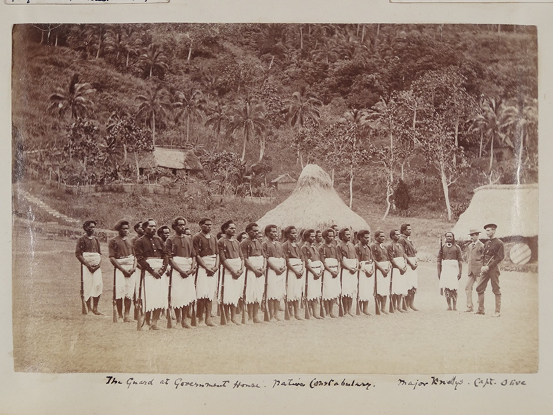 Sepia photograph o a group of men in white barkcloth clothing, standing orderly in front of a group of three commanders.  A house and dense forest is visible behind them.