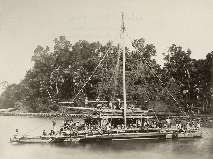 A sepia photograph of a drum, with many people on board. Trees are visible on the bank of the river. Writing can be seen through the reverse of the photograph.