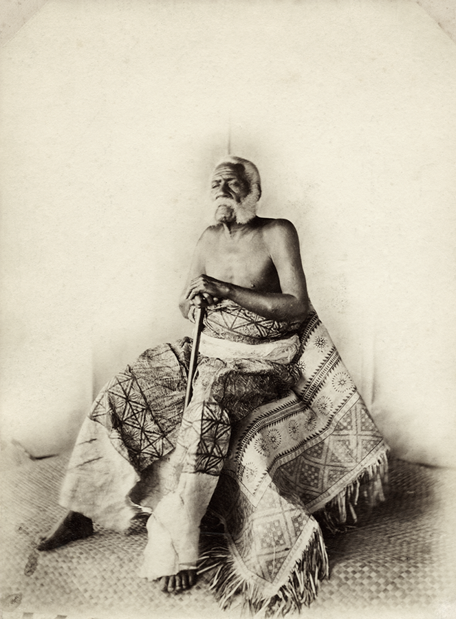 Sepia photograph of a man, seen full length, dressed in a ceremonial skirt piece and carrying a staff. He has a white beard and hair.