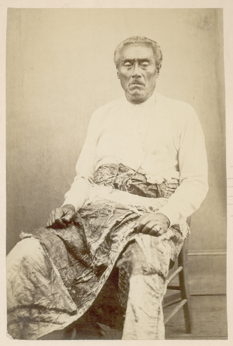 Sepia photograph of a man wearing a white long sleeve top and a ceremonial barkcloth, seated on a stool. He has short hair and looks directly at the camera.