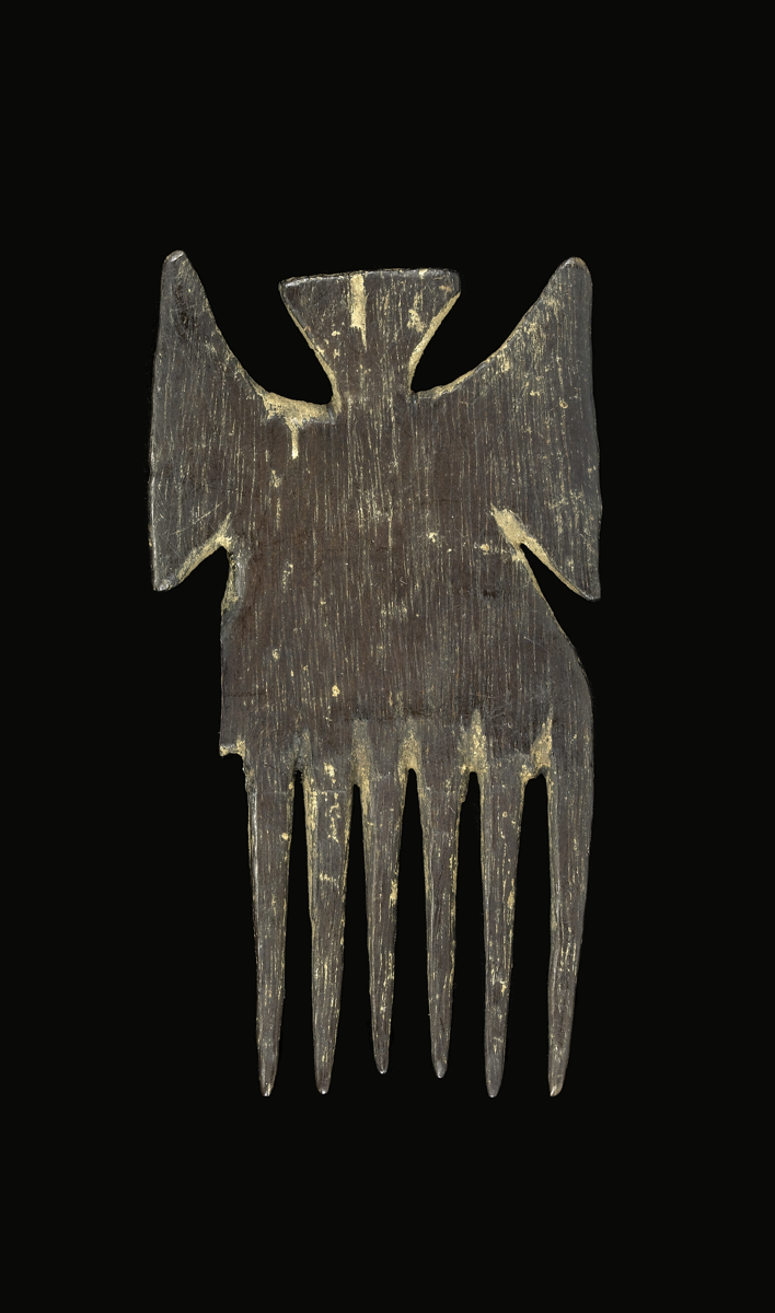 A comb carved from wood. There are six sub-cylindrical and tapering prongs with one prong broken away and missing. The handle consists of two triangular projections at the side and one on the top formed by cut-outs around them. The wood is stained dark br