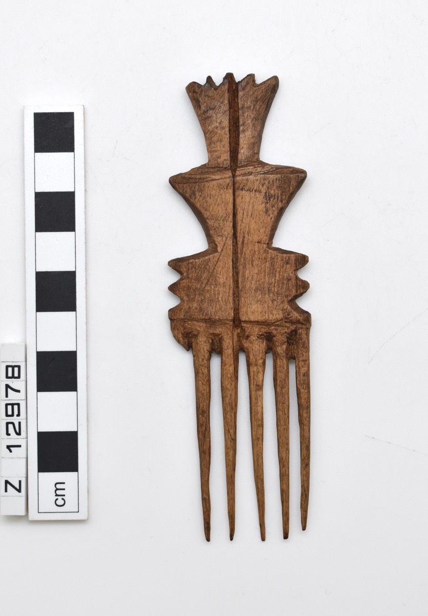 A comb carved from wood with six cylindrical and tapering prongs, one of which has broken away and is missing. The handle of the comb has cut-out sections at each side forming triangular shapes and serrated edges.