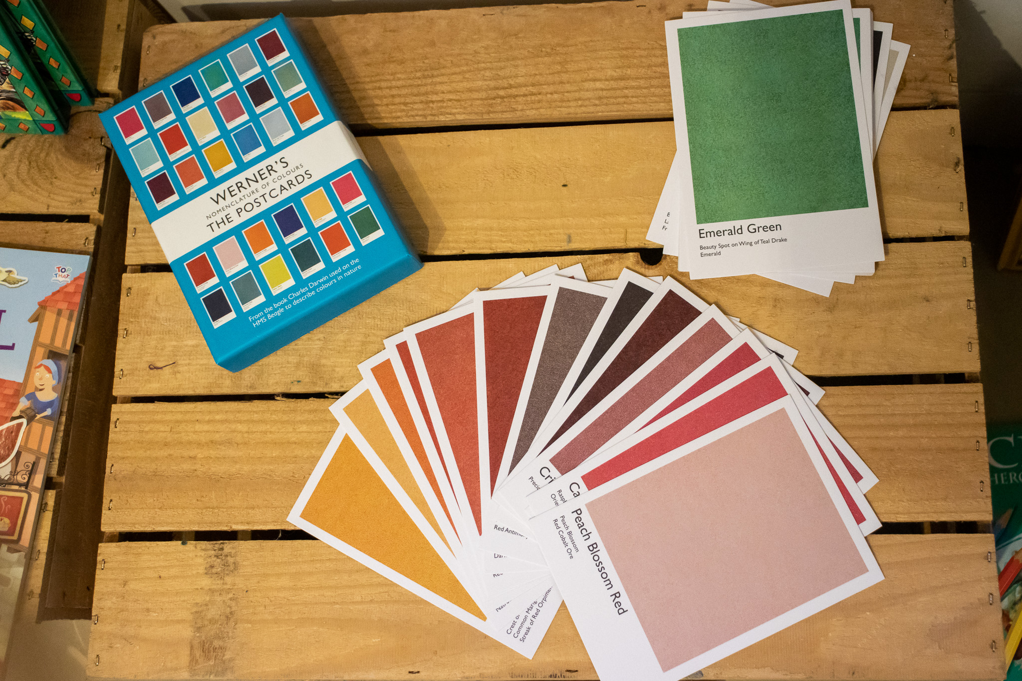Display of postcards from 'Werner's Nomenclature of Colours: The Postcards'. A fanned-out display showing the colourful cards, each with a different colour, and most visible are 'peach blossom red' and 'emerald green'. Displayed on top of a wooden box.