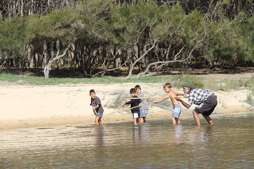 Rod Mason crouching with a spear, showing four young boys how to fish, in shallow waters near a beach.