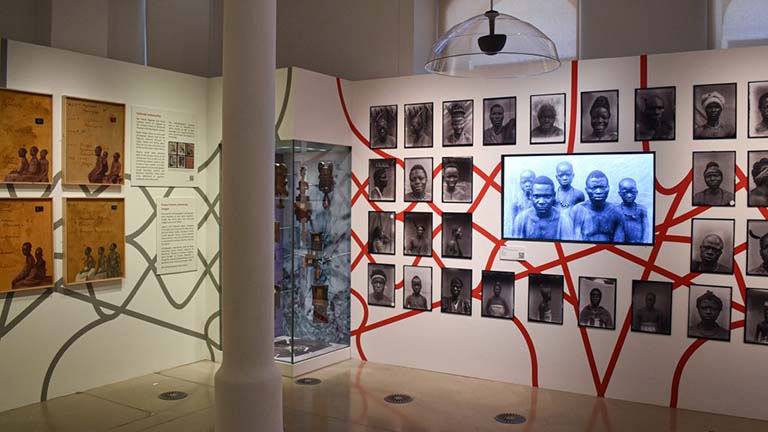An exhibition shot of the Re Entanglements exhibition. On the right there is a display of headshots/photographs of people, with a video seen in the centre, intertwined by a graphic of red lines.