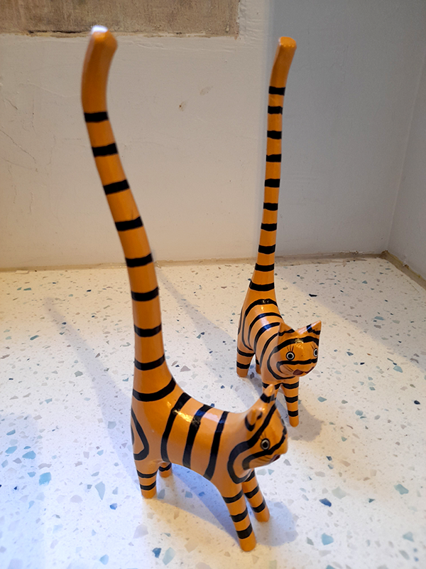 Two ring holders in the shape of a cat, orange with black stripes, with their tails the ring holder segments. Sat against an MAA shop counter.