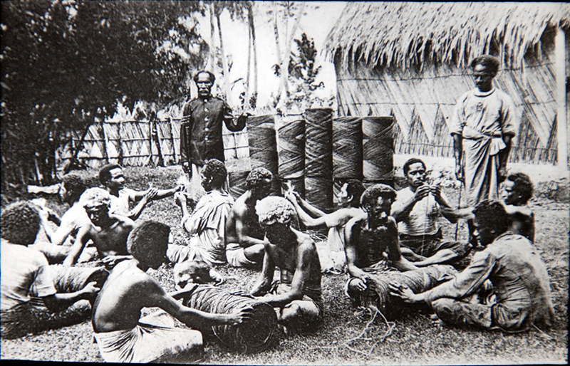 Men sit in front of coir coils, at work rolling more. Two men stand behind, overseeing the work.