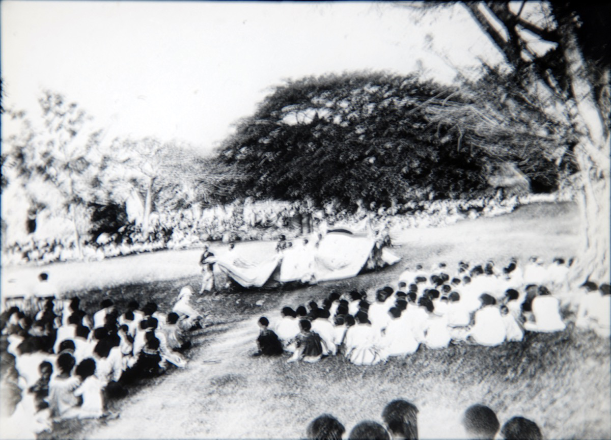 Slightly blurry black and white photograph of a ceremony, with many people sitting around a central performance