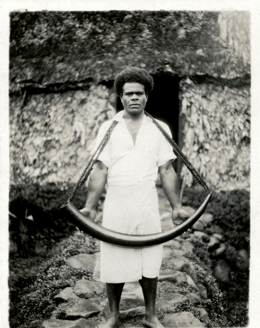 Sepia photograph of a man in white clothing holding an elephant tusk with a long cord.