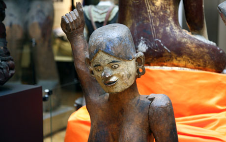 A wooden statue, life sized, of the wife of a chief of a village. Visible in the gallery with objects behind it.