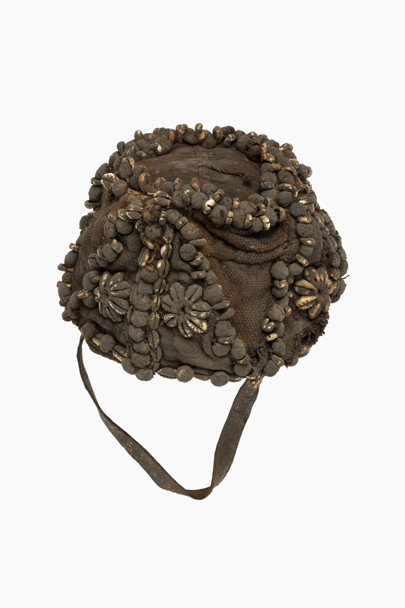  hat in woven cloth with a chin strap attached to the edges. Six flowers made from cowrie shells decorate the sides of the hat