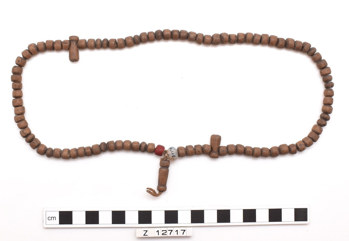 Necklace of wooden beads with two glass beads. Most beads are cylindrical, with the centre point having extra beads to make a feature at the front. Two long cylindrical beads are spaced around the necklace.