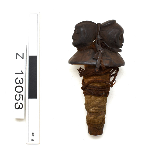 Bottle stopper with two back to back heads. Leather and fibre strings wrapped around the object. Dark brown.