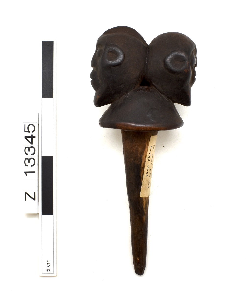 Bottle stopper with rounded top and two back to back heads. Faces are oval shaped with long chins; mouths are in whistling position. Dark brown.