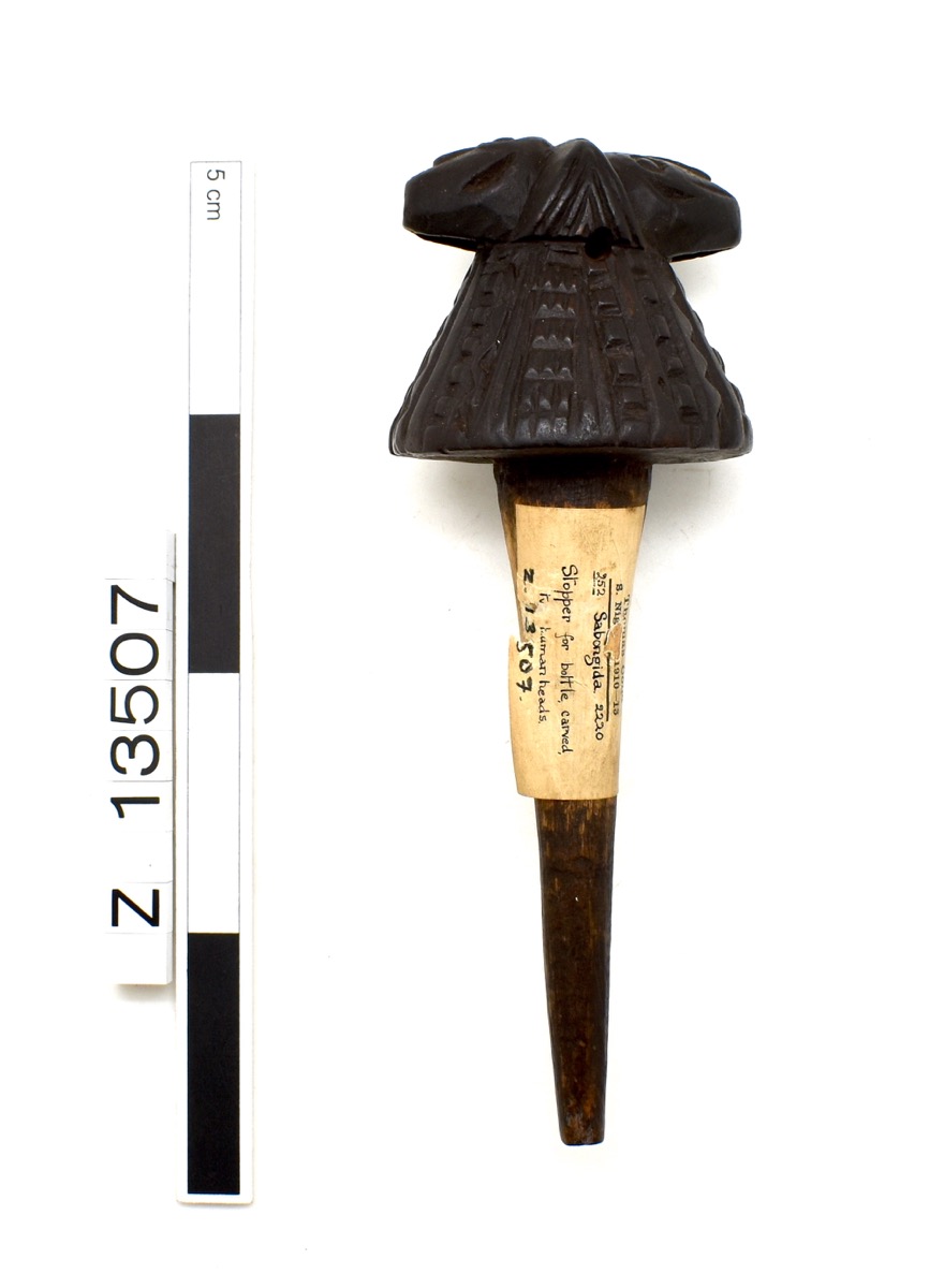 Bottle stopper with a larger rounded top and two heads, back to back, resting atop it. Heads are possibly animal but it is hard to tell from the photo. The rest of the top features geometric design. A sticker is attached with accession information.
