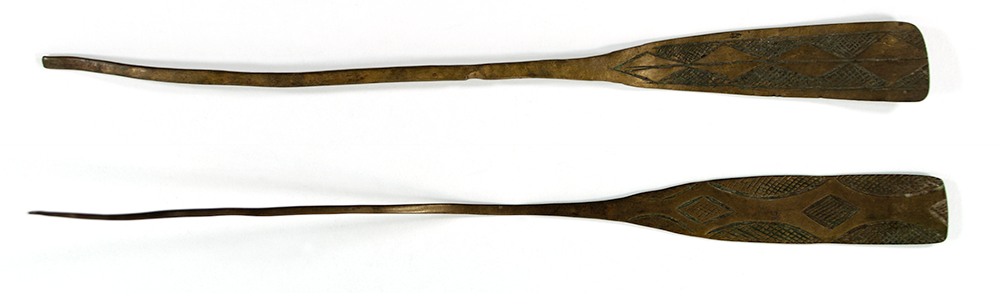 Two brass hairpins. One with a flat, rectangular head and geometric head pattern, and the other with a flat rectangular head tapering in towards the pin. 