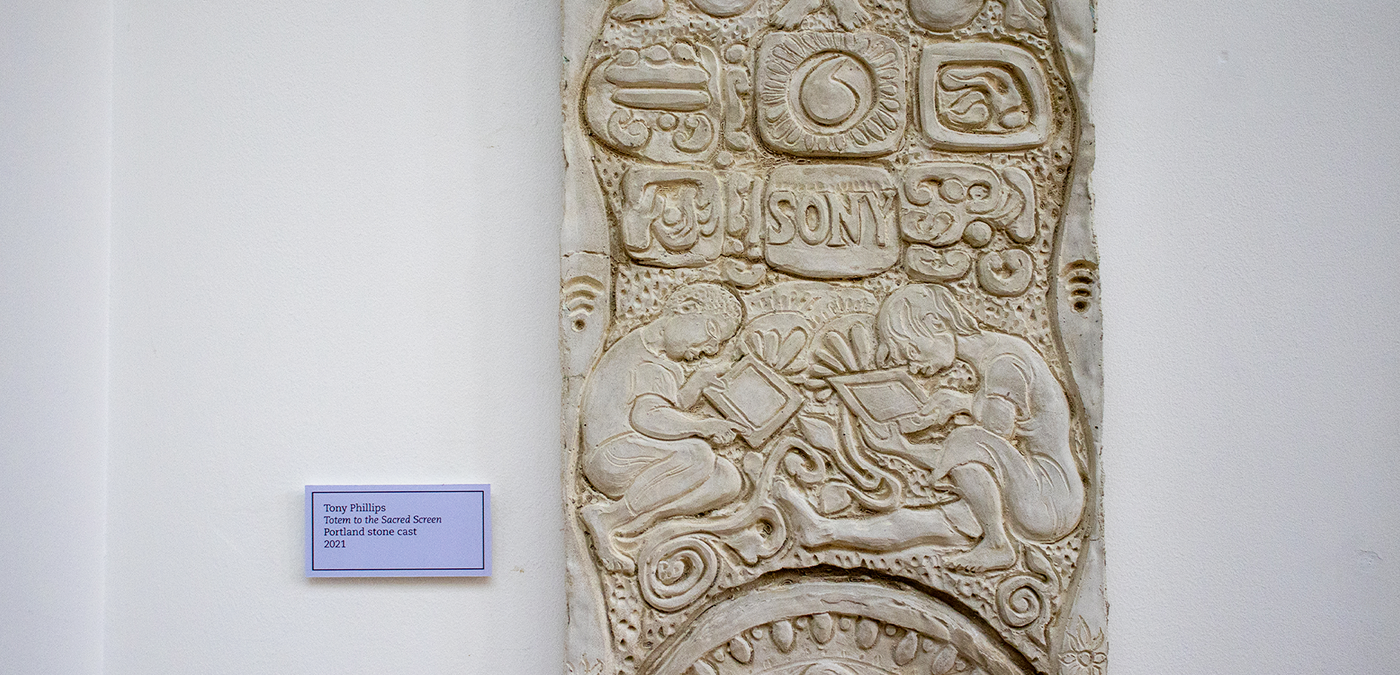 Close up of 'Totem to the Sacred Screen', a carving by Tony Phillips with modern logos (wifi, Sony, and more) and in the centre two children crouching over tablets.