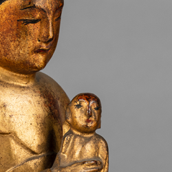 Close-up of a gilt wooden statue of Guanuin with an infant (Songzi Guanyin). Bright gold statue, against a grey background.