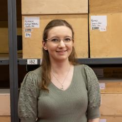Staff photograph of Lizzy Peneycad, smiling at the camera. She wears a green top and glasses, and has brown hair; she is standing in front of a selection of boxes on shelves, labelled varieties of 'Roman'.