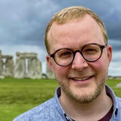 A man with fair hair and glasses stands outside with Stonehenge in the background.