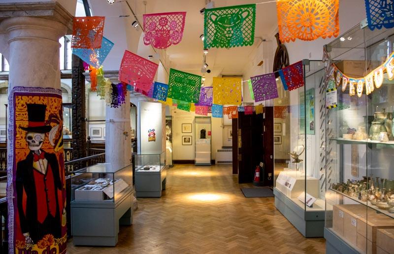 A long thin museum gallery decorated to celebrate Día de Muertos (Day of the Dead). There is brightly coloured bunting strung across the gallery and a depiction of a skeleton dressed in a three-piece suit and top hat attached to a column on the left.
