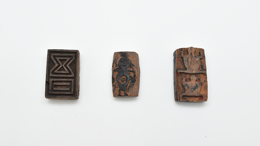 Three body stamps in a horizontal row. L-R: a rectangular stamp with geometric raised relief, in a rectangle and hourglass shape; a rounded stamp with raised relief of a human figure, the raised part black from ink; a rectangular stamp with raised reliefs